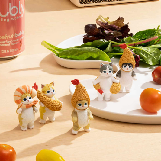 A collection of small PVC figurines of cats in various colors, designed to resemble fried shrimp, displayed on a table with some vegetables.