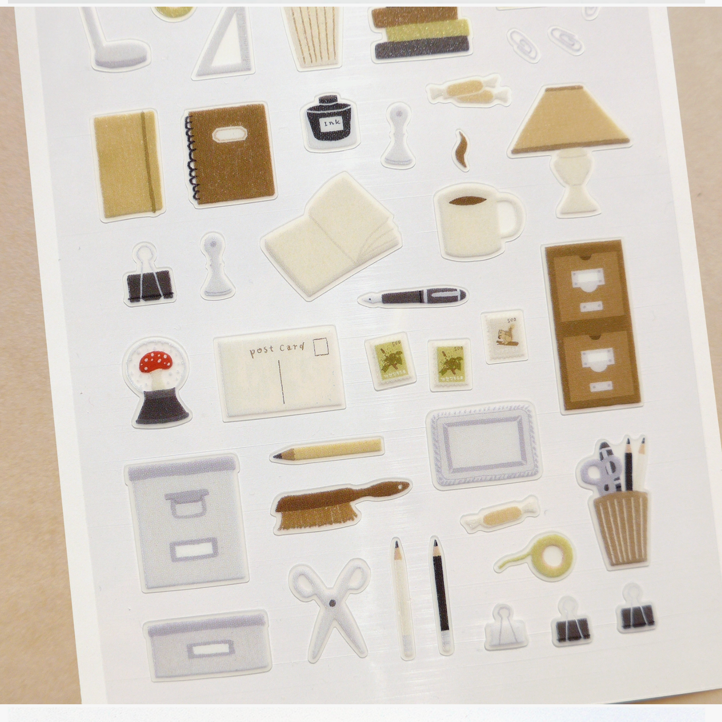 A sheet of small desk and office-themed stickers on a white backing, photographed from above on a beige surface. The stickers include items such as folders, pens, scissors, pencils, a coffee cup, books, clips, and small accessories in neutral beige, gray, black, white, and brown tones.