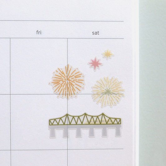 A close-up of a printed calendar or planner page with columns labeled fri and sat, featuring simple line drawings of fireworks and a bridge in muted yellow, pink, green, and gray tones.