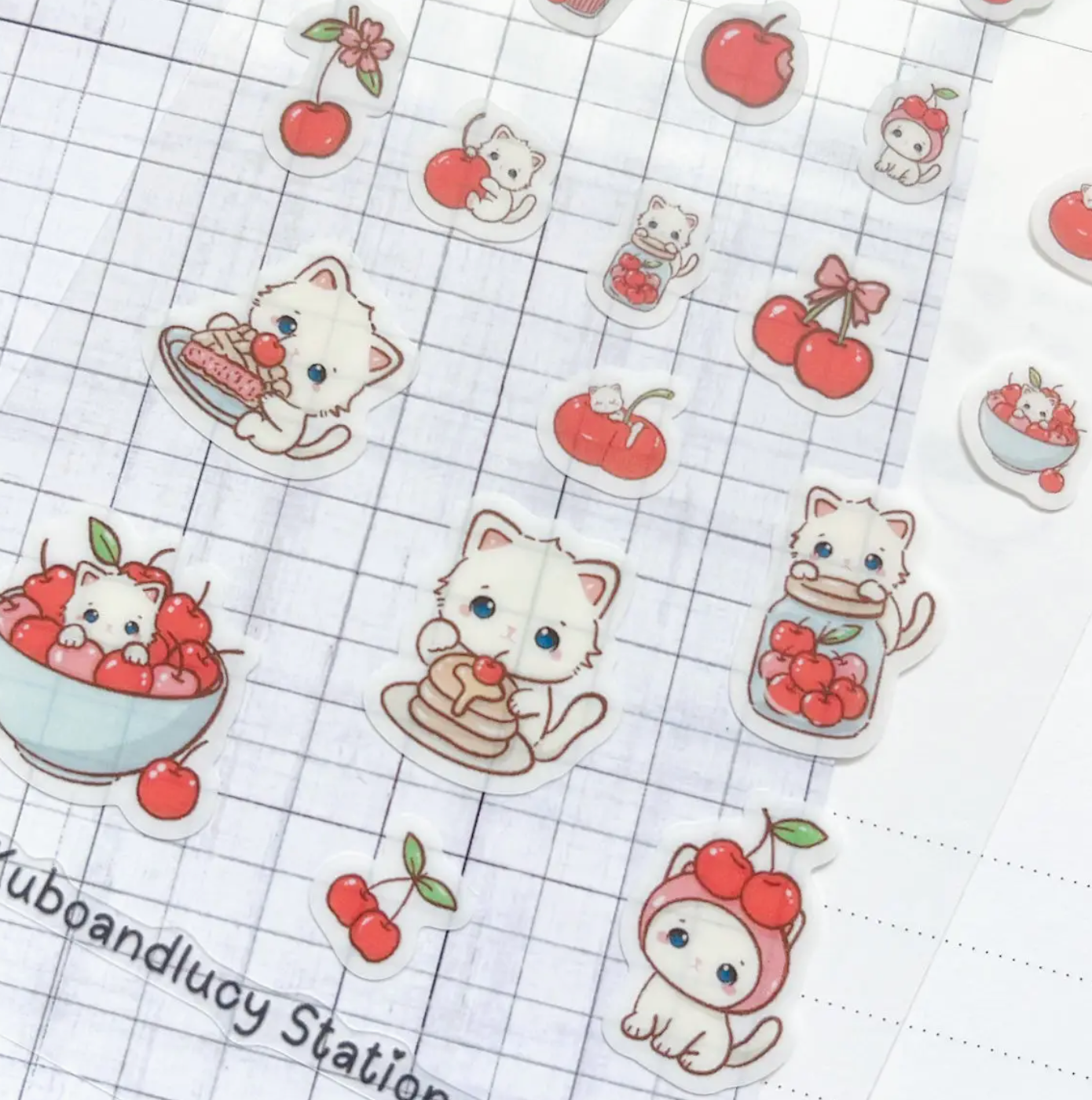 A sheet of cute cartoon sticker decals featuring white cats, red cherries, and desserts, laid out on a white grid paper background.