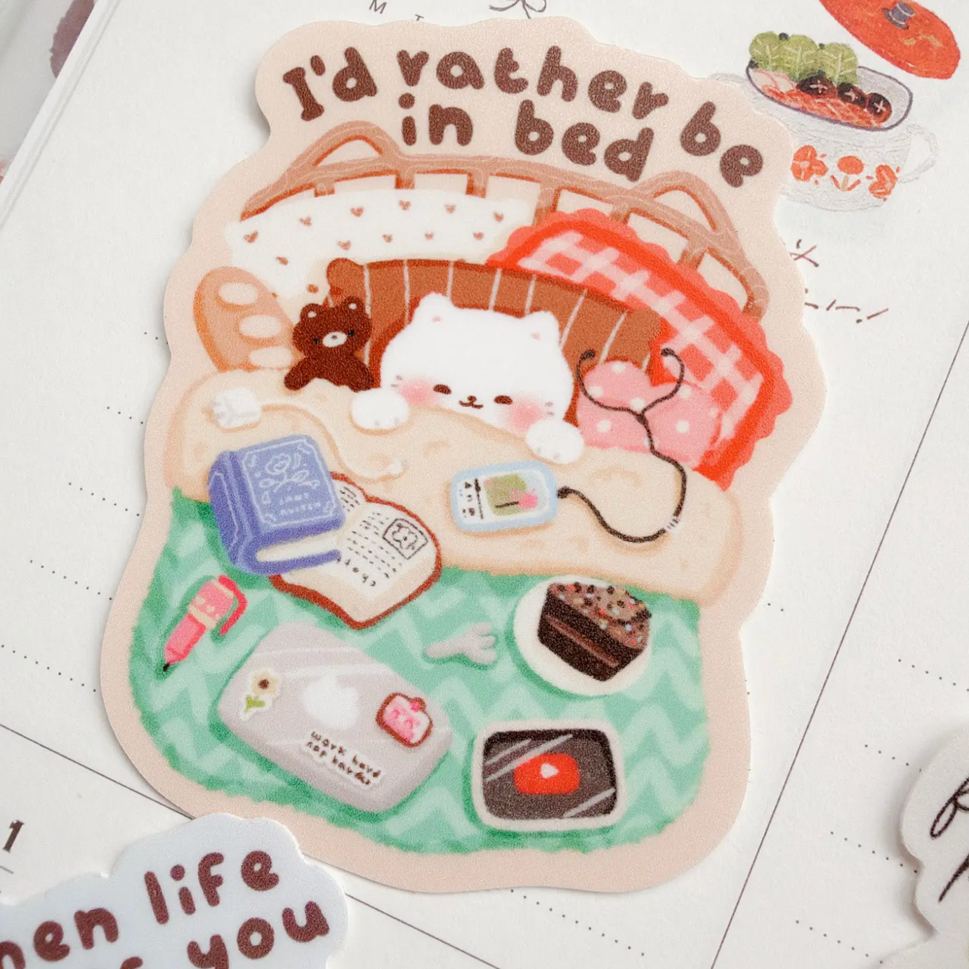 A die-cut sticker featuring a cute illustration of a white cat sleeping in a basket with a red plaid blanket, surrounded by small daily items like a book, phone, cake, and cupcake, with the text 'I'd rather be in bed'. The sticker is displayed on a lined notebook page.