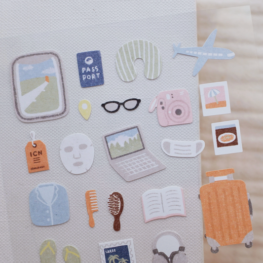 A set of cute travel-themed sticker decals arranged on a textured white sheet, featuring illustrations of a suitcase, passport, plane, mask, glasses, camera, laptop, and other travel accessories.