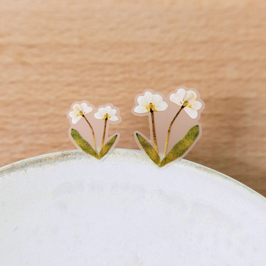 A pair of small floral stud earrings with white flowers, yellow centers, and green leaves on a translucent pink backing, displayed on the edge of a white ceramic dish against a light wood background.