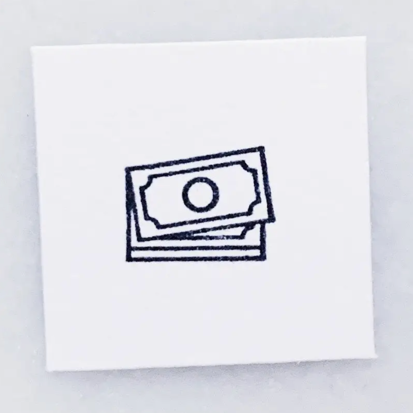 A square white card or sticker featuring a simple black line drawing of stacked dollar bills, centered on a light gray background.