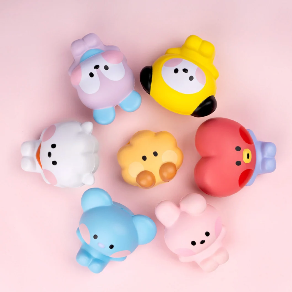 Six colorful cartoon character squishy toys arranged in a circle on a soft pink background. The toys include pastel purple, yellow, white, yellow-orange, red, light blue, and light pink characters, each with cute cartoon faces and simple body shapes.