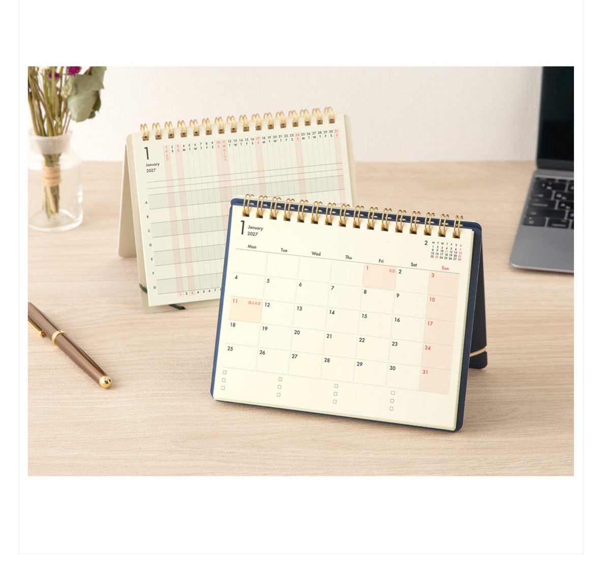 A spiral-bound desk calendar shown open on a light wood tabletop, with a second calendar standing behind it. The front calendar has a navy blue cover and an off-white page, with a few highlighted dates in soft pink.
