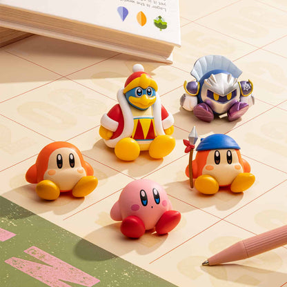 A collection of colorful Kirby blind box figures inspired by Kirby, including characters resembling Kirby, King Dedede, Waddle Dee, Meta Knight, and Bandana Waddle Dee, seated on a desk next to a book and a pen.