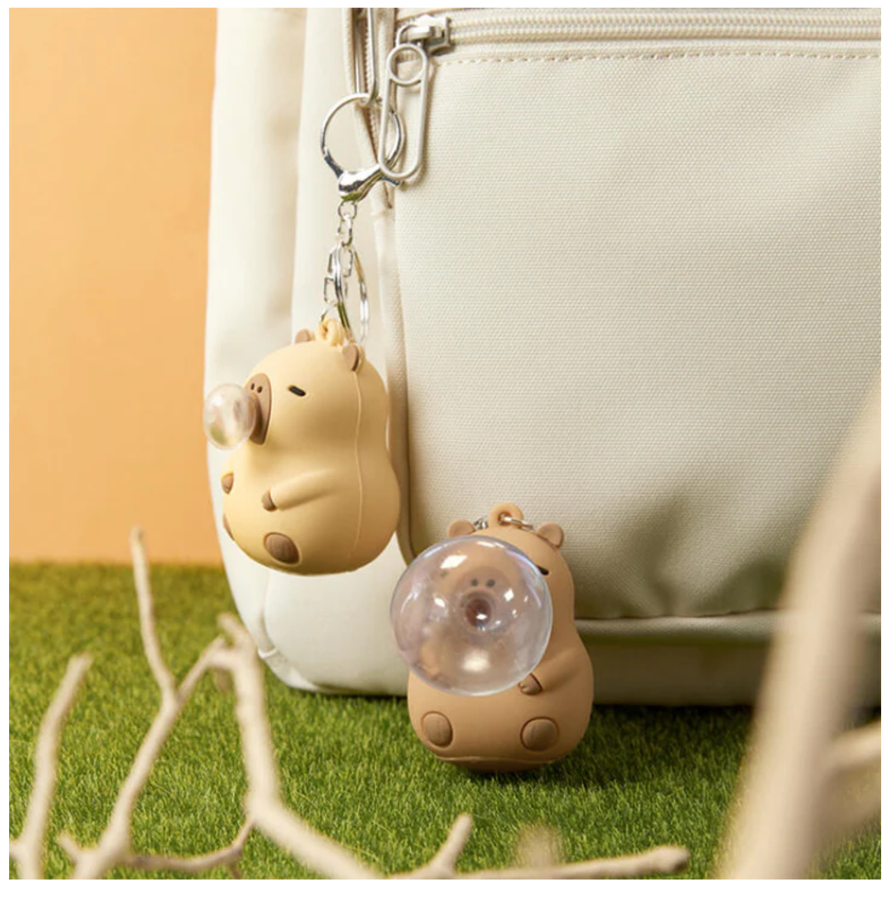 Two capybara keychains with one hanging on a bag and the other sitting next to bag on a beige back ground and grass. 