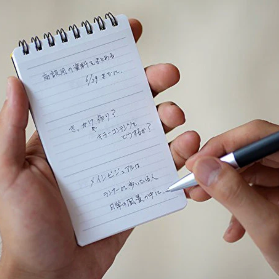 Hand holding a small spiral-bound notepad with handwritten text and a pen.