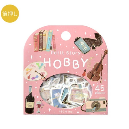 A pink retail package labeled 'Petit Story HOBBY' containing 45 decorative sticker pieces. The package illustration includes books, a film reel, a cello, a paint palette, a wine bottle, and a camera, with a clear window showing the assorted stickers inside.