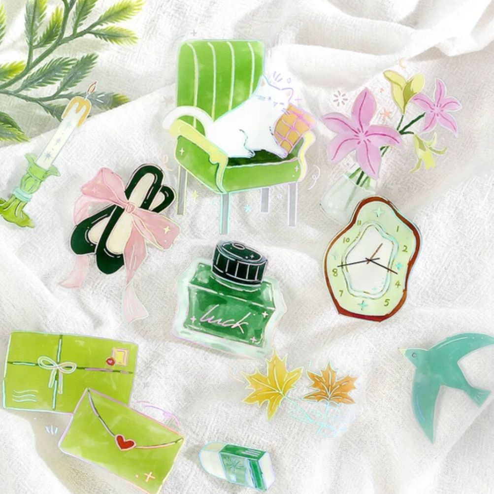 A collection of cute, hand-drawn style watercolor stickers arranged on a textured white fabric background. The stickers feature a green armchair with a white cat, pink flowers, a green ink bottle with the brand name 'luck', green shoes, a green gift box, a green envelope with a red heart, a green clock, and other small whimsical illustrations.