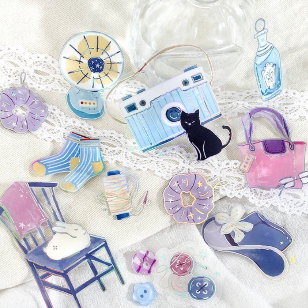 A collection of cute, pastel-colored watercolor-style enamel pins arranged on a white lace fabric background. The pins include a light blue camera with a black cat, pink and purple handbags, blue striped socks, a chair with a bunny, a small fan, and other whimsical accessories.