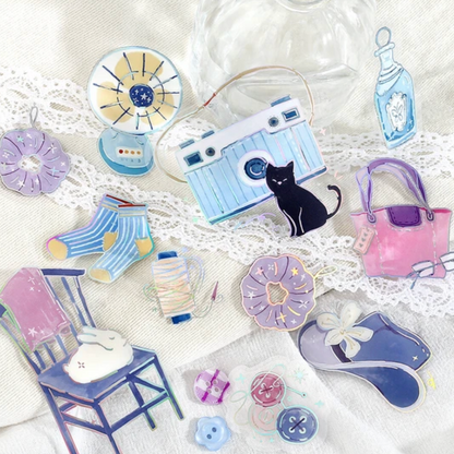A collection of cute, pastel-colored watercolor-style enamel pins arranged on a white lace fabric background. The pins include a light blue camera with a black cat, pink and purple handbags, blue striped socks, a chair with a bunny, a small fan, and other whimsical accessories.