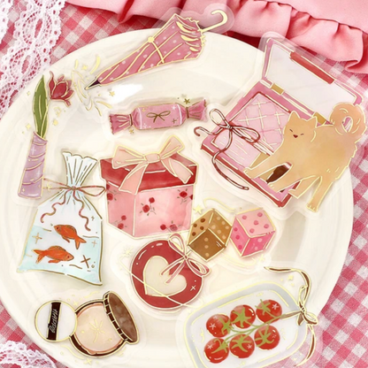 A collection of cute, pastel pink and red enamel pins styled on a white plate with a pink and white checkered cloth background. The pins feature whimsical designs including a cat, umbrellas, gift boxes, fish, cherries, and makeup items.