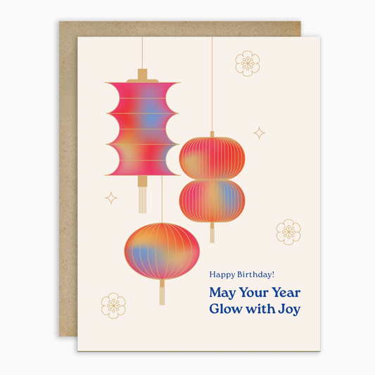 A birthday greeting card with a kraft envelope, featuring three colorful hanging lantern illustrations in pink, orange, and blue on an off-white background, with the text 'Happy Birthday! May Your Year Glow with Joy'.
