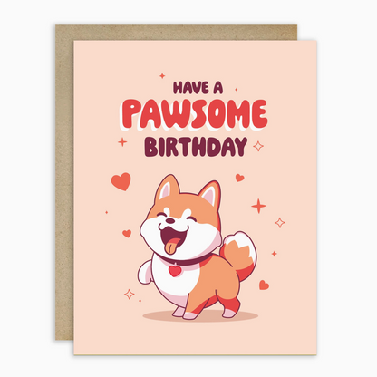 A cute birthday greeting card with a kraft envelope behind it. The card has a light peach background with the text 'HAVE A PAWSOME BIRTHDAY' and an illustration of a smiling Shiba Inu dog wearing a heart collar.