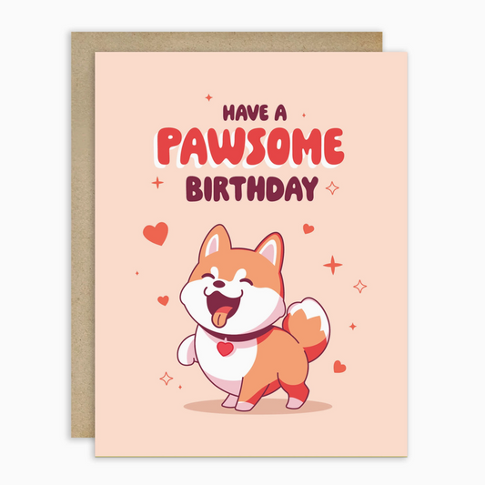 A cute birthday greeting card with a kraft envelope behind it. The card has a light peach background with the text 'HAVE A PAWSOME BIRTHDAY' and an illustration of a smiling Shiba Inu dog wearing a heart collar.