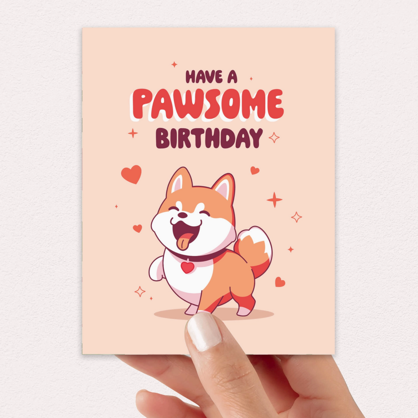 A hand holds a square birthday card with a peach background, featuring the text 'HAVE A PAWSOME BIRTHDAY' above a cartoon Shiba Inu dog with a red heart-shaped tongue. The card is photographed against a light neutral wall.