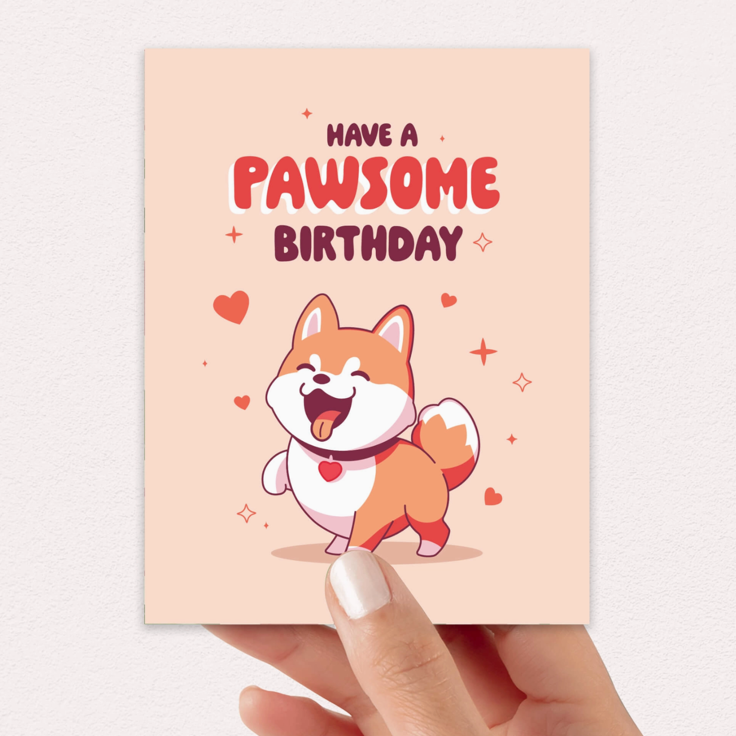 A hand holds a square birthday card with a peach background, featuring the text 'HAVE A PAWSOME BIRTHDAY' above a cartoon Shiba Inu dog with a red heart-shaped tongue. The card is photographed against a light neutral wall.