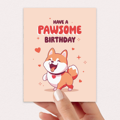 A hand holds a square birthday card with a peach background, featuring the text 'HAVE A PAWSOME BIRTHDAY' above a cartoon Shiba Inu dog with a red heart-shaped tongue. The card is photographed against a light neutral wall.