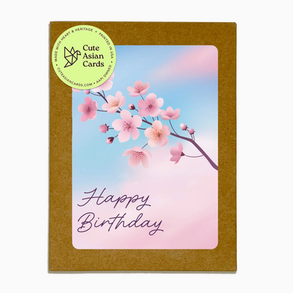 A square Happy Birthday card with a brown kraft paper border and a central illustration of pink cherry blossoms against a soft blue and pink gradient background. The words 'Happy Birthday' are written in purple script.