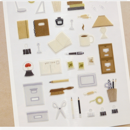 A sheet of small desk and office-themed stickers on a white backing, photographed from above on a beige surface. The stickers include items such as folders, pens, scissors, pencils, a coffee cup, books, clips, and small accessories in neutral beige, gray, black, white, and brown tones.