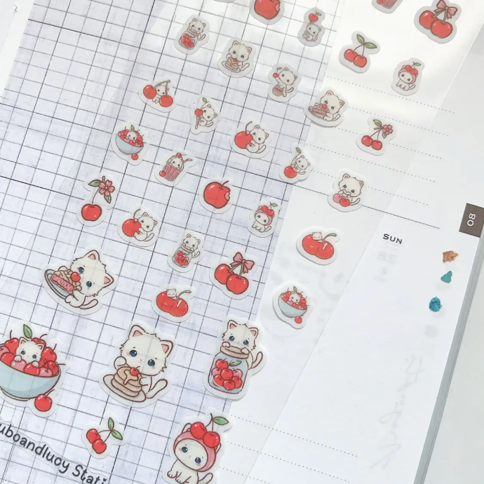 A close-up of cute planner sticker sheets featuring kawaii cats, cherries, and small food illustrations arranged on a white grid planner page.