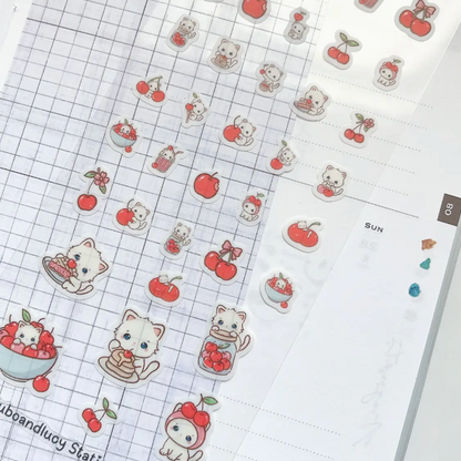 A close-up of cute planner sticker sheets featuring kawaii cats, cherries, and small food illustrations arranged on a white grid planner page.