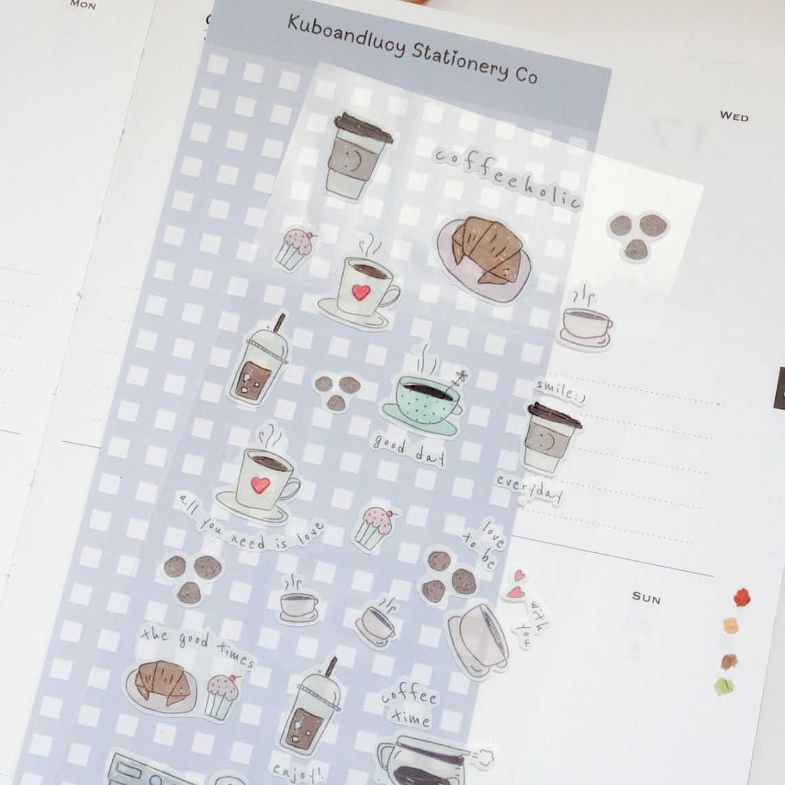 A sheet of cute coffee-themed planner stickers by Kubaandlucy Stationery Co, featuring illustrations of takeaway coffee cups, croissants, cupcakes, and handwritten text like 'coffee addict' and 'good day!' on a light blue and white gingham background.