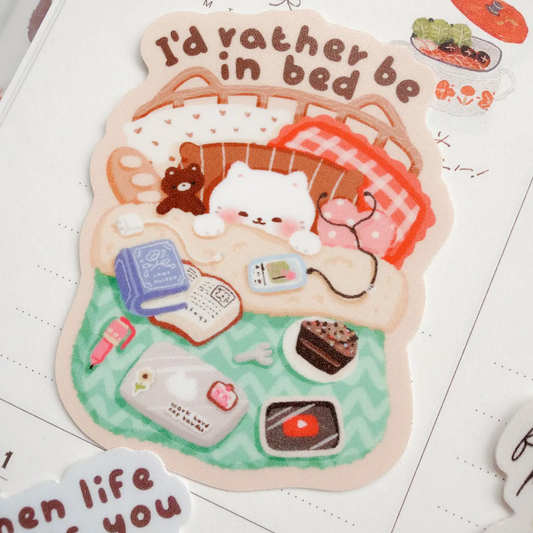 A die-cut sticker featuring a cute illustration of a white cat sleeping in a basket with a red plaid blanket, surrounded by small daily items like a book, phone, cake, and cupcake, with the text 'I'd rather be in bed'. The sticker is displayed on a lined notebook page.