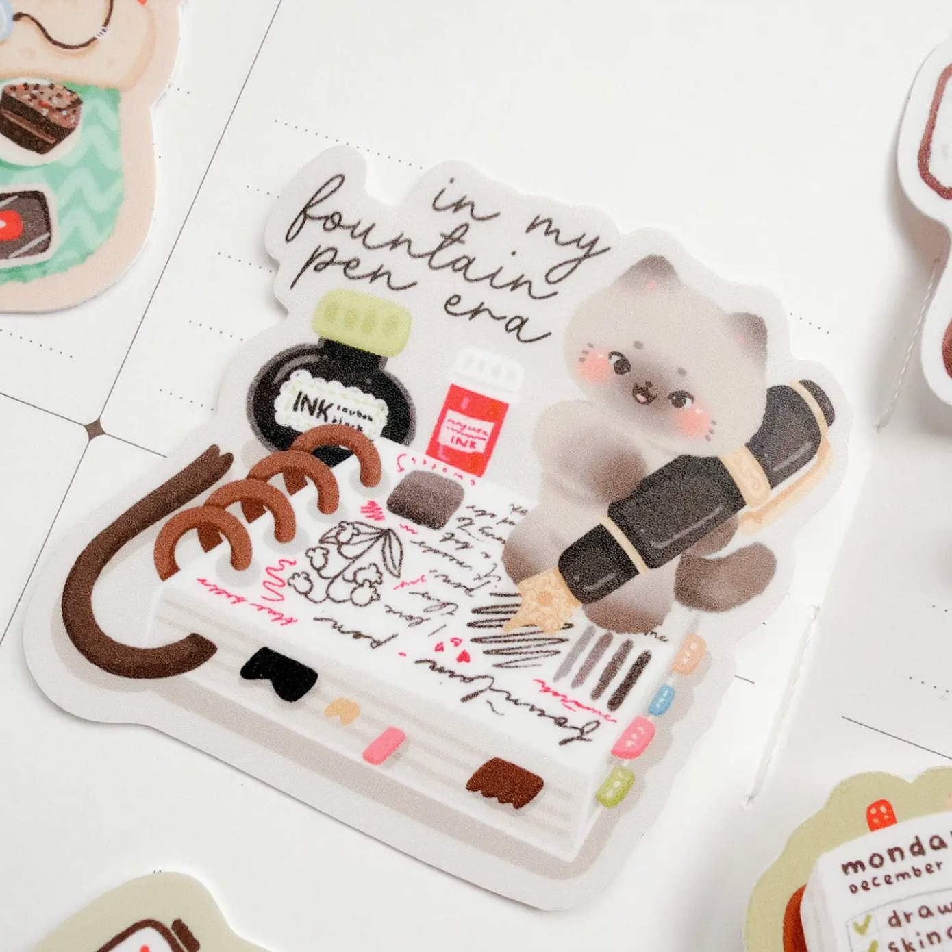 A sheet of cute planner stickers featuring a fluffy gray kitten holding a black and gold pen, surrounded by stationery doodles, handwritten notes, and small pastel accents, displayed on a white grid paper background.