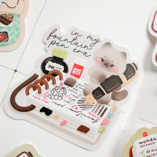 A sheet of cute planner stickers featuring a fluffy gray kitten holding a black and gold pen, surrounded by stationery doodles, handwritten notes, and small pastel accents, displayed on a white grid paper background.