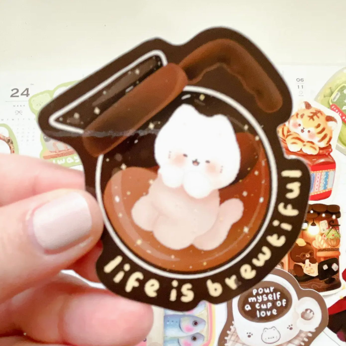 A hand with light pink nail polish holds a die-cut sticker featuring a cute white cat sitting in a cup of coffee, surrounded by the text 'life is beautiful'. The sticker has a dark brown border and a warm brown coffee background with small gold star details.