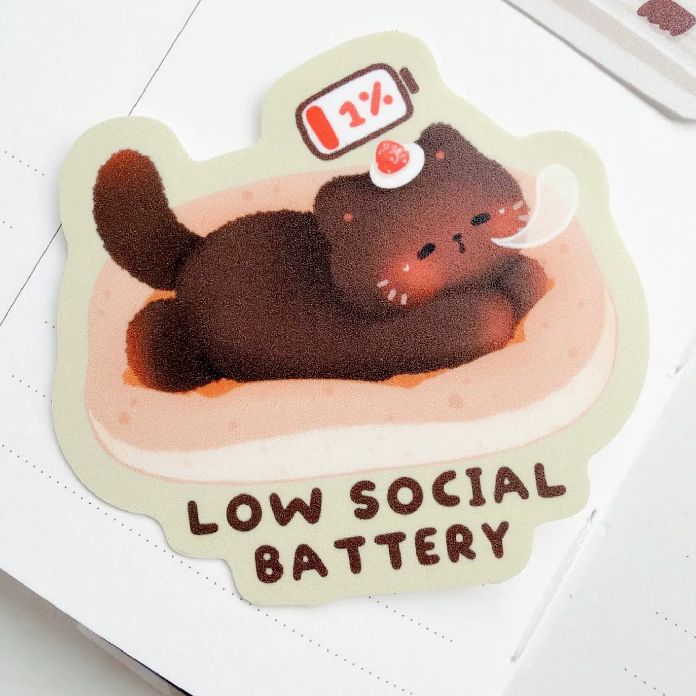 A die-cut sticker featuring a cute cartoon cat lying in a bed with a 1% battery icon above it and the text 'LOW SOCIAL BATTERY' at the bottom, shown on a white notebook page.