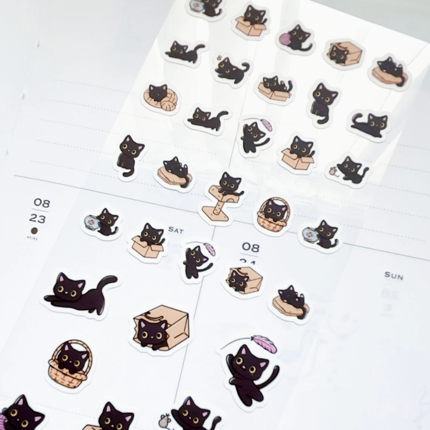 A sheet of cute black cat planner stickers scattered on a white planner page, featuring various playful poses with boxes, baskets, and small accessories.