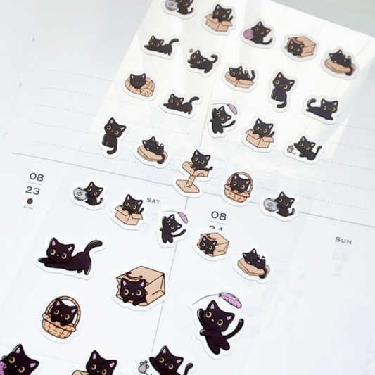 A sheet of cute black cat planner stickers scattered on a white planner page, featuring various playful poses with boxes, baskets, and small accessories.