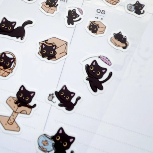 A sheet of cute cartoon black cat stickers displayed on a white lined planner page. The stickers depict various playful poses of black cats, including sitting, lying down, playing with a feather, using a cat tree, and holding a fish bowl.