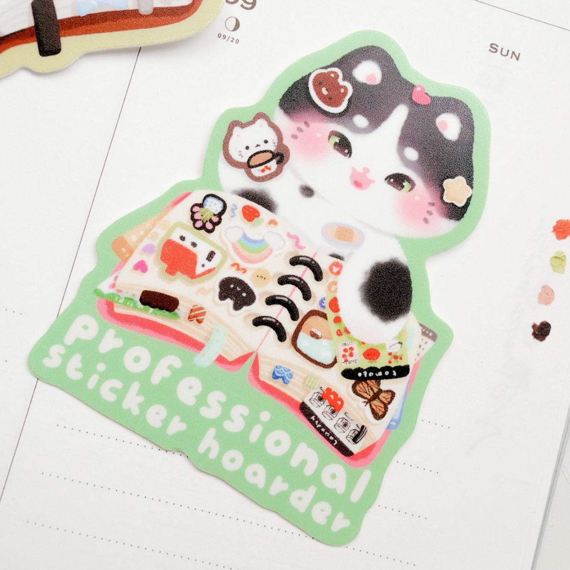 A cute die-cut sticker featuring a black-and-white cat character surrounded by stickers and the text 'professional sticker hoarder' on a pale green background, placed on a white planner page.
