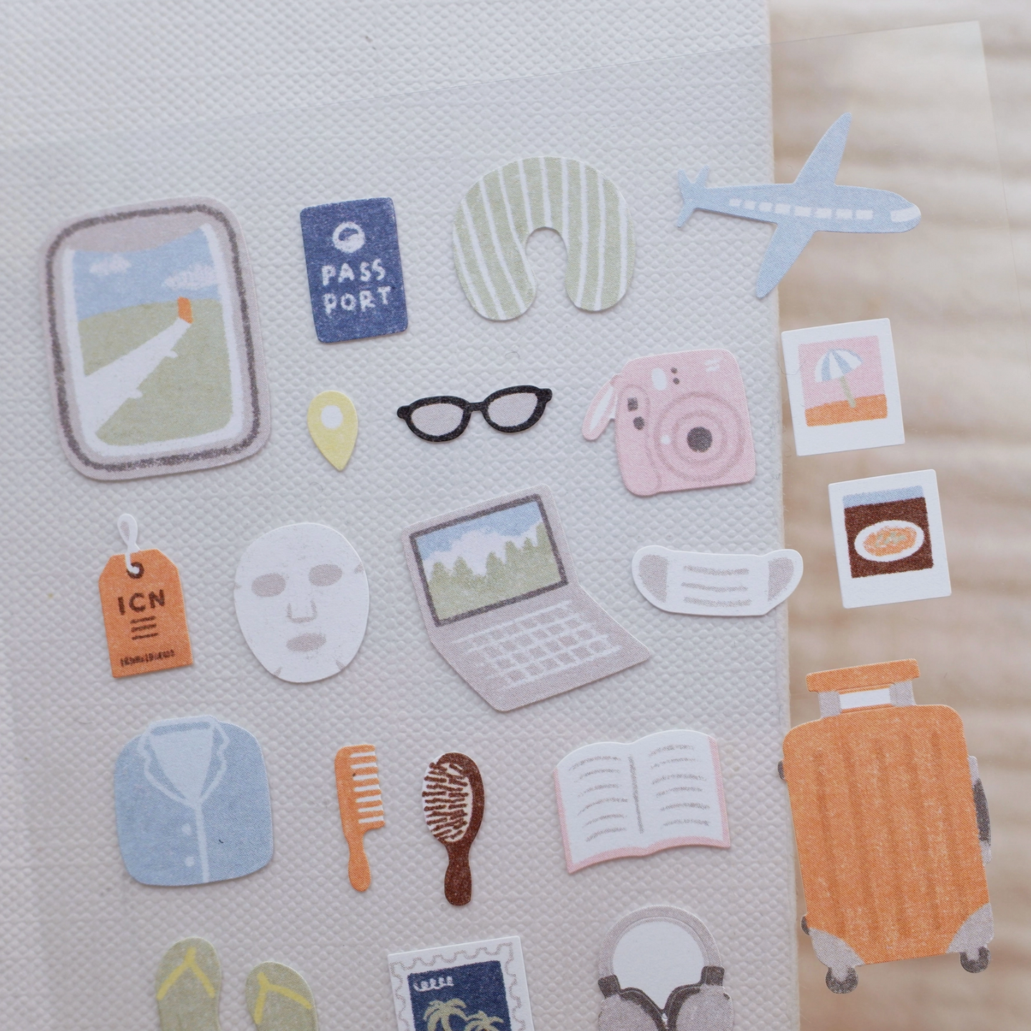 A set of cute travel-themed sticker decals arranged on a textured white sheet, featuring illustrations of a suitcase, passport, plane, mask, glasses, camera, laptop, and other travel accessories.