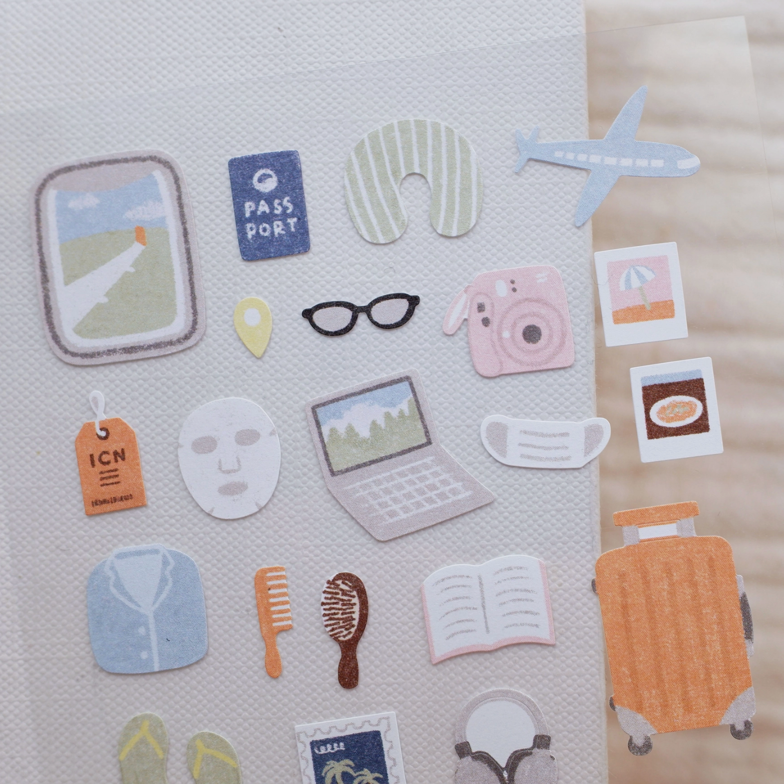 A set of cute travel-themed sticker decals arranged on a textured white sheet, featuring illustrations of a suitcase, passport, plane, mask, glasses, camera, laptop, and other travel accessories.