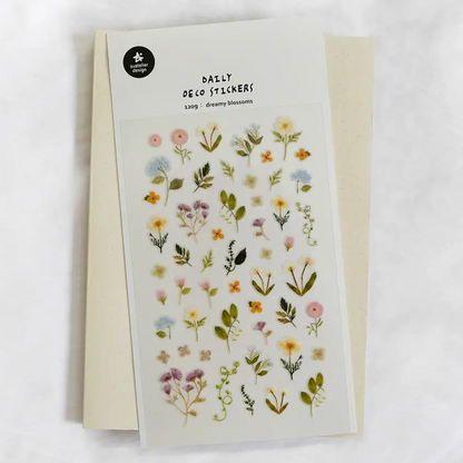 A sheet of floral deco stickers with small pressed flowers and green leaves on a white backing, placed over a cream envelope on a light marble surface. The sheet reads 'DAILY DECO STICKERS' and shows the brand name 'The Paper Flower' in a black circle at the top left.