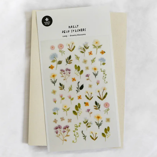 A sheet of floral deco stickers with small pressed flowers and green leaves on a white backing, placed over a cream envelope on a light marble surface. The sheet reads 'DAILY DECO STICKERS' and shows the brand name 'The Paper Flower' in a black circle at the top left.