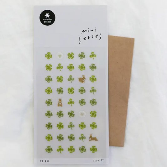 A sheet of small decorative stickers labeled 'mini series' with mostly green clover motifs and a few brown animal shapes, placed on a white background with a brown envelope behind it.