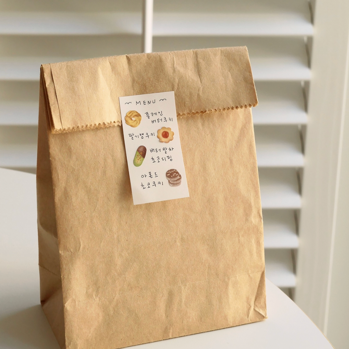 A kraft paper bag with a small handwritten label, standing on a white surface in front of white window shutters.