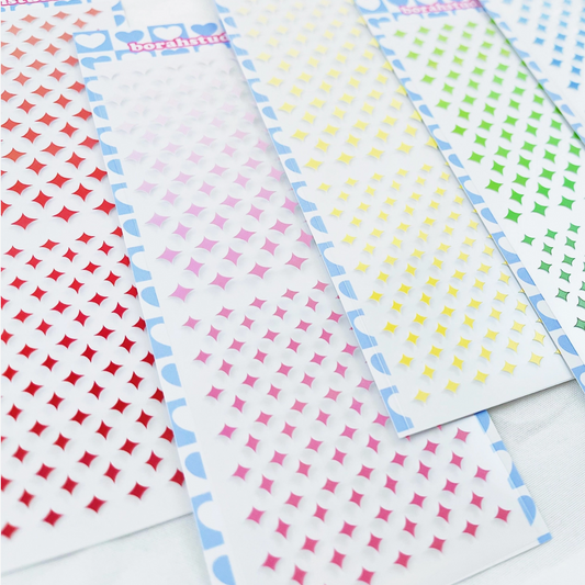 Several narrow strips of decorative stickers or labels are arranged diagonally on a white surface. Each strip has a repeating pattern of small diamond/star-like shapes in different colors: red, pink, yellow, green, blue, and white.