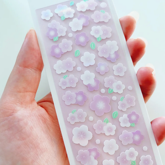A hand holding a long, narrow clear plastic strip decorated with small pastel cherry blossom stickers in lavender, white, and mint green.