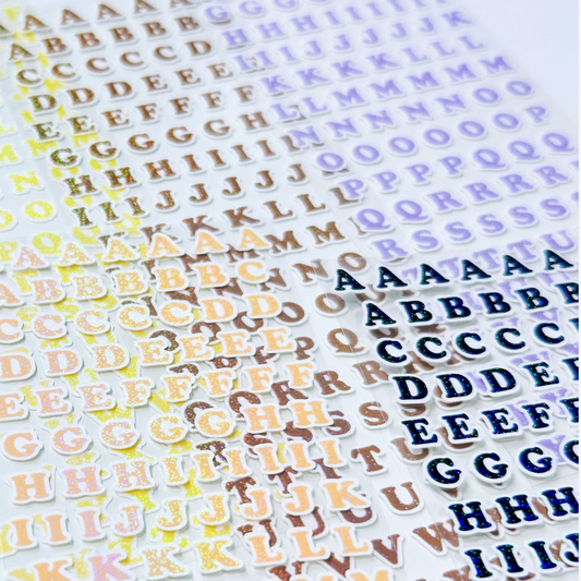A close-up of multiple sheets of die-cut alphabet stickers featuring uppercase letters in various colors, including gold, brown, blue, purple, green, and pink, arranged diagonally on a white background.