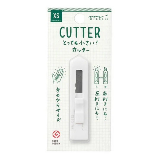 A white handheld paper cutter with a gray blade window, mounted on a white grid-patterned retail card with green text and illustrations. The card shows the brand MIDORI and size XS.