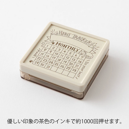A square monthly habit tracker stamp with a cream-colored top and a clear base, featuring a printed calendar grid and Japanese text below on a white background.