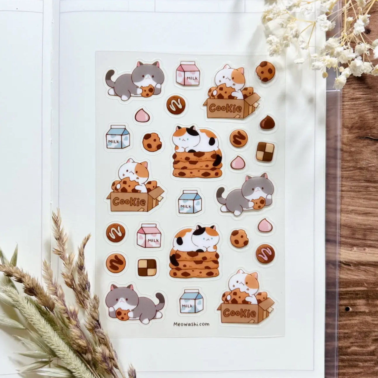 A sheet of cute cat-themed stickers displayed on an open notebook, surrounded by dried flowers on a rustic wooden surface. The stickers feature gray and orange tabby cats playing with milk boxes, cookies, chocolate bars, and hearts.